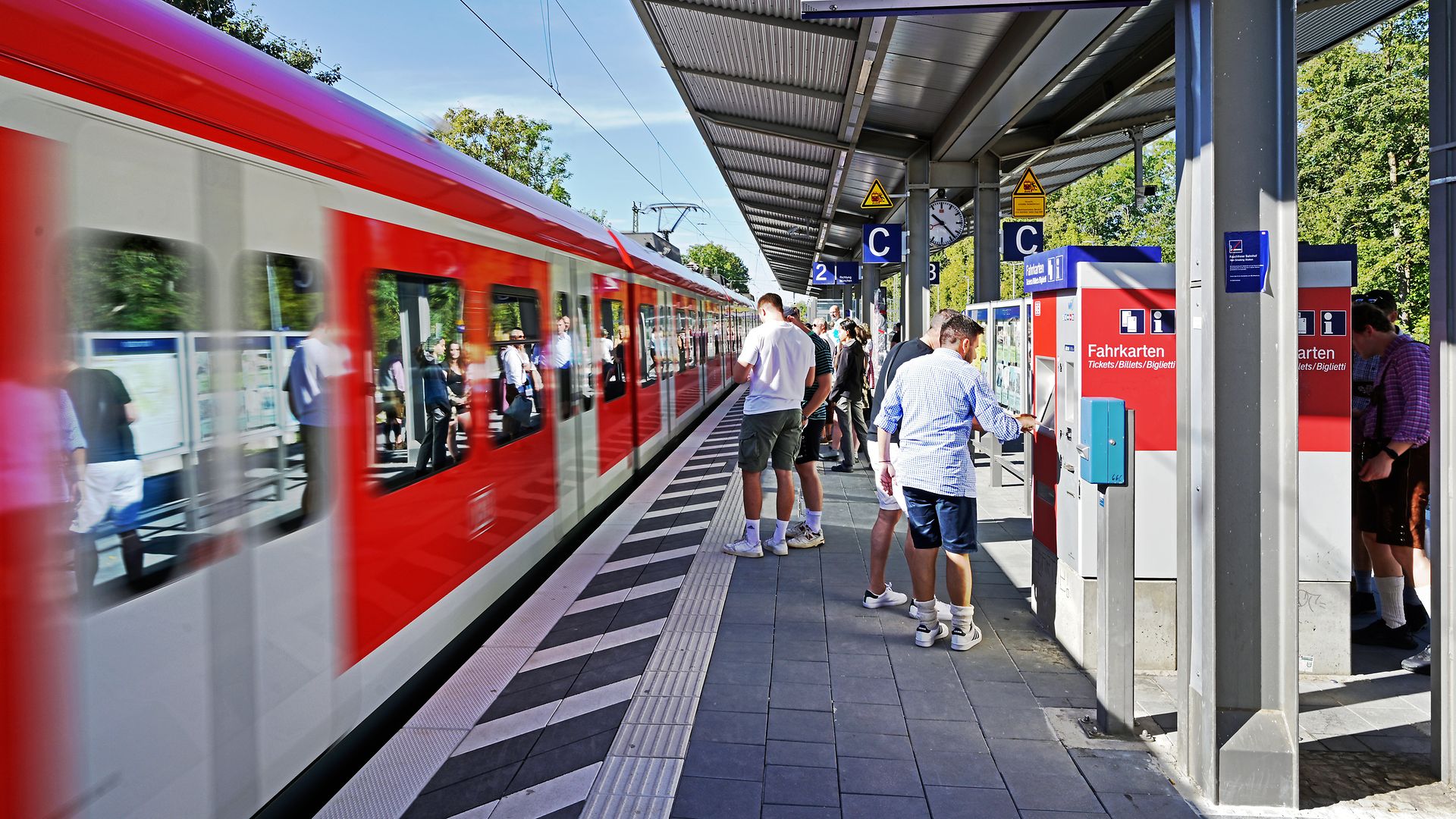 Reisende warten hinter dem taktilen Blindenleitstreifen auf den einfahrenden Zug am Bahnsteig